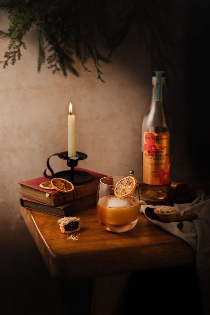 Moody festive cocktail styled with a mince pie garnish, candlelight and vintage books, featuring The Reverend Hubert liqueur in a cinematic winter setting. Atmospheric drinks photography by Lou Carruthers, Cotswolds Food and Hospitality Photographer