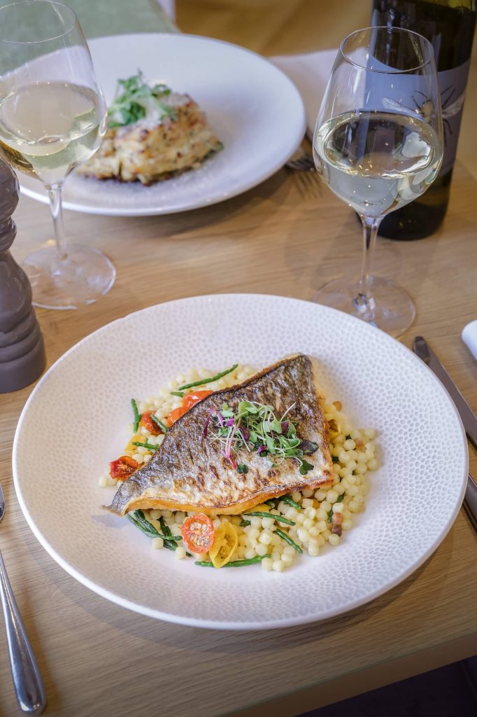 Pan-seared fish served with seasonal vegetables and grains, styled with white wine on a softly lit restaurant table at Siddington Park, photographed by Lou Carruthers, Lou Carruthers Photography, Hospitality & Drinks Photographer, Cotswolds and Menorca