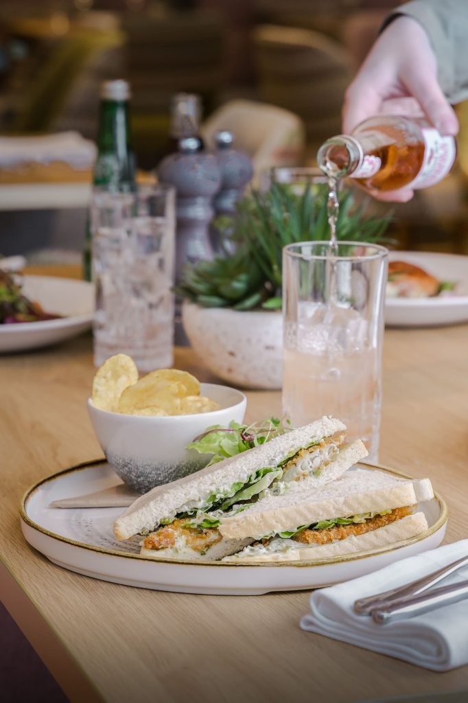 Casual lunch setting with freshly prepared sandwiches, crisps and drinks being poured at the table inside Siddington Park, photographed by Lou Carruthers, Lou Carruthers Photography, Hospitality & Drinks Photographer, Cotswolds and Menorca