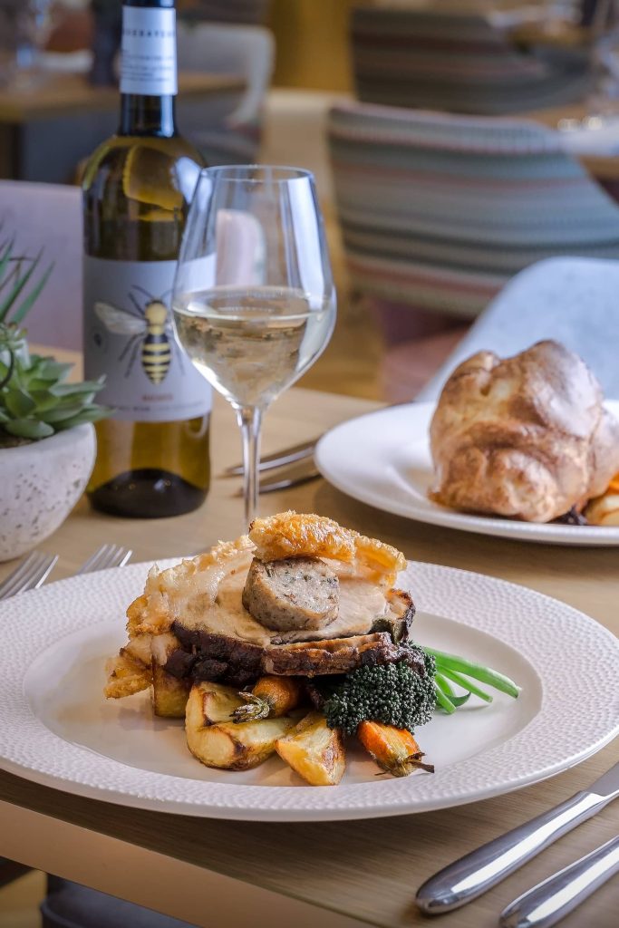 Seasonal roast pork served with golden roast potatoes, vegetables and a glass of white wine on a restaurant table at Siddington Park, photographed by Lou Carruthers, Lou Carruthers Photography, Hospitality & Drinks Photographer, Cotswolds and Menorca