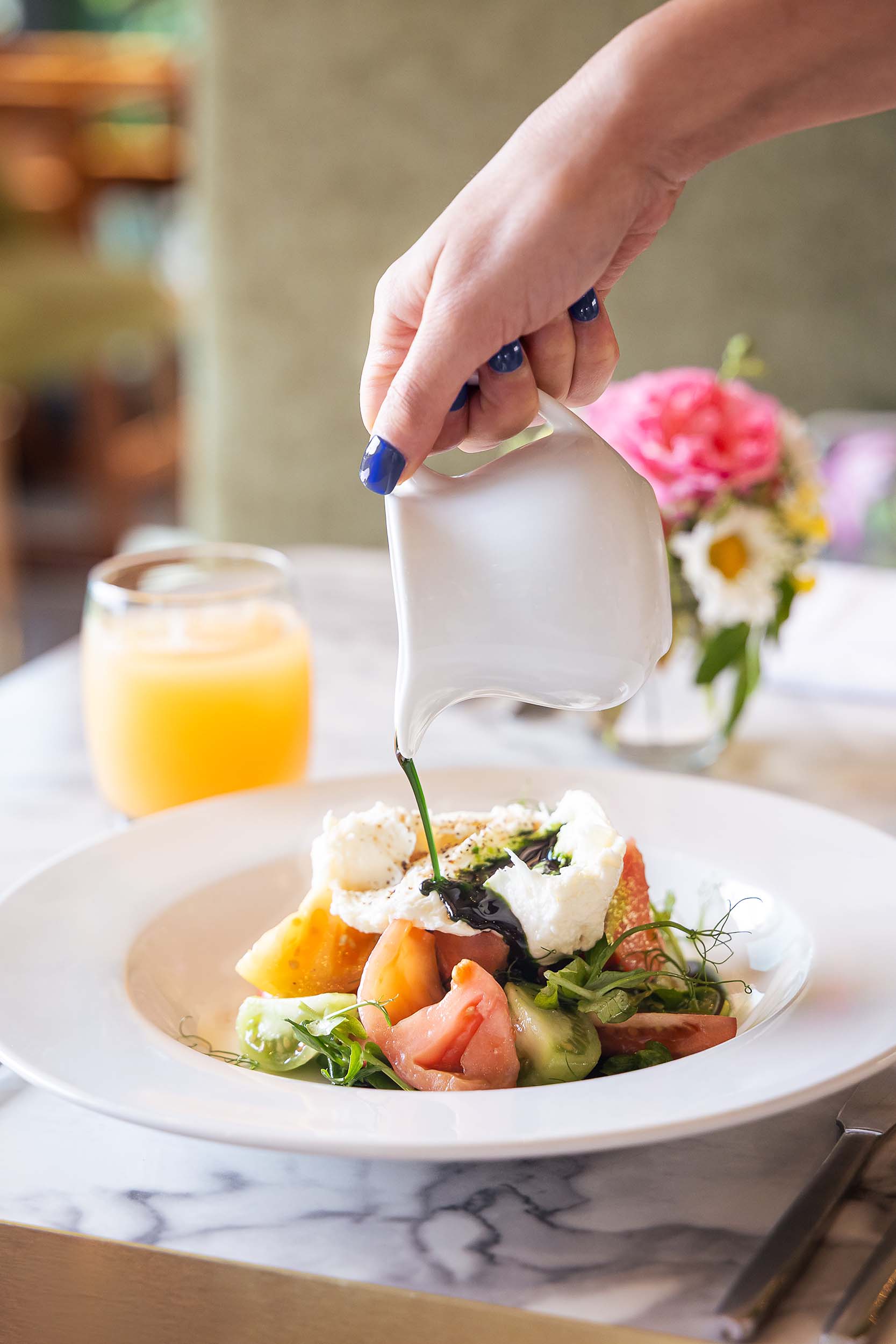 A hand pours balsamic glaze over a burrata salad with tomatoes and fresh greens on a restaurant table.