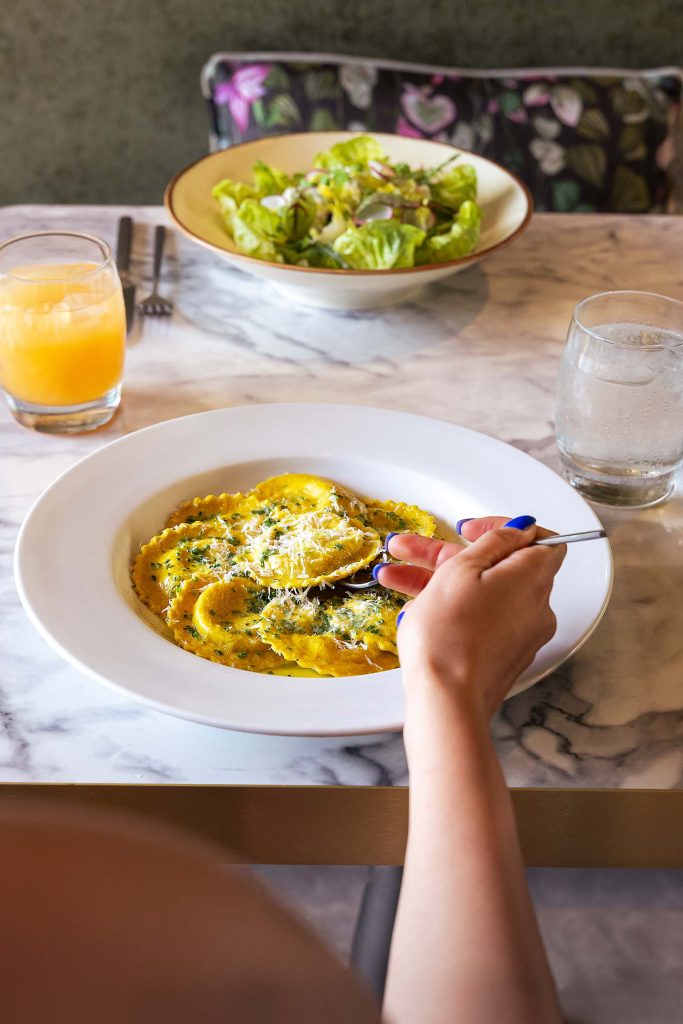 A diner lifts a forkful of ravioli from a bowl of pasta at a restaurant table set with drinks and salad.