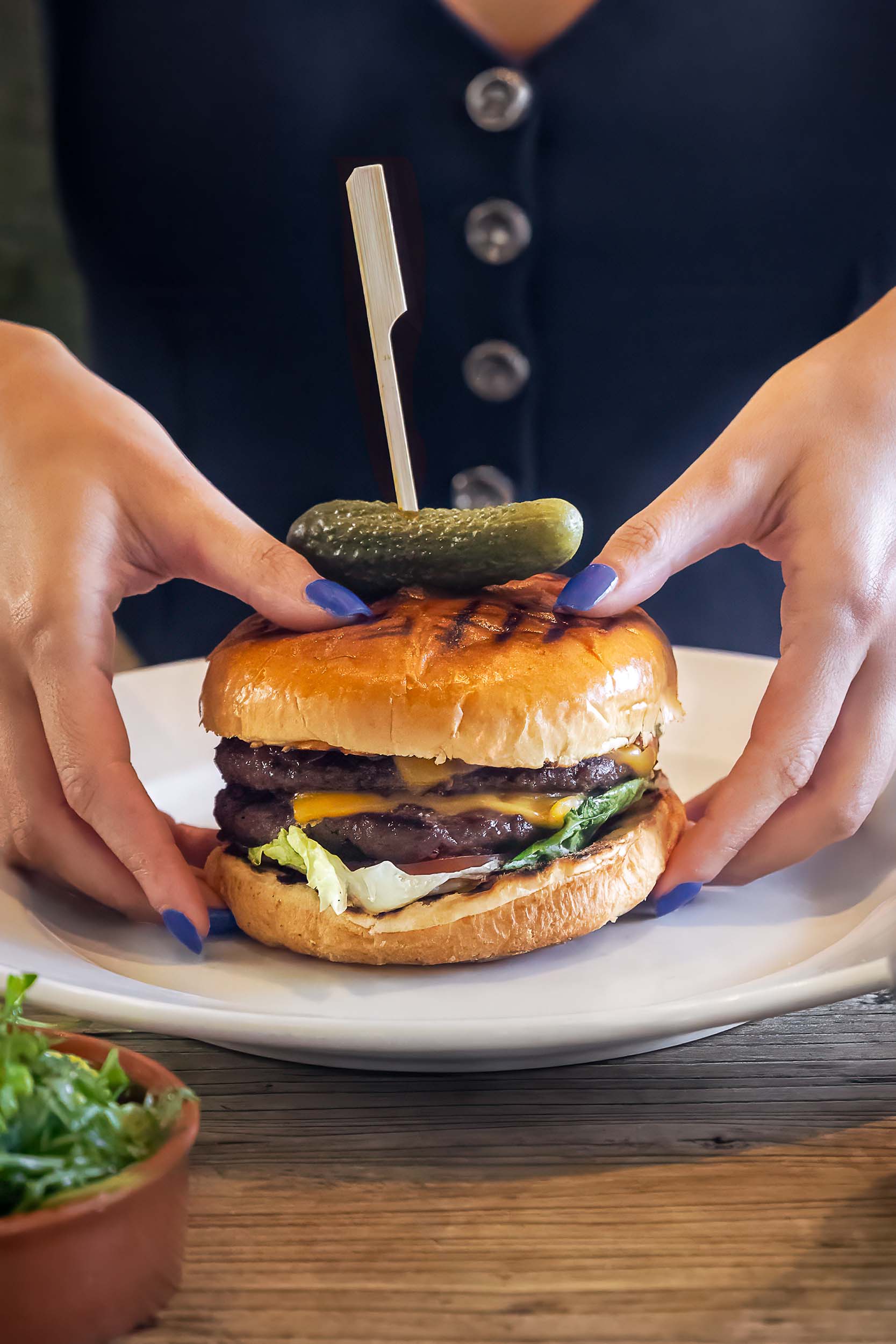 A gourmet cheeseburger is presented on a white plate as a diner positions it before eating.